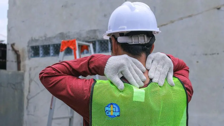 A construction worker wearing a hard hat, reflective vest, and gloves, is holding their neck with their hands.