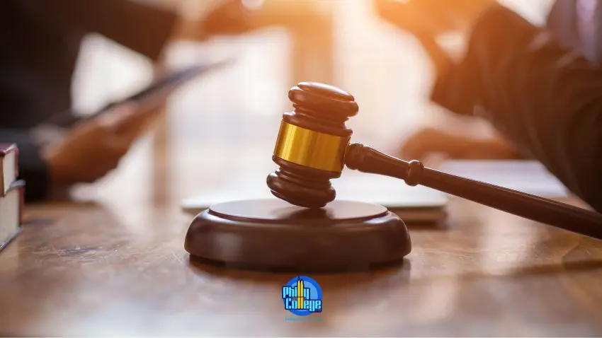 A close-up shot of a wooden gavel resting on its base on a wooden table, with blurred figures in the background and warm sunlight filtering light.