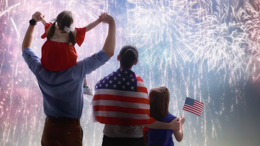 A group of people looking at fireworks, one holding an American flag, and another person has an American flag draped over their shoulders.