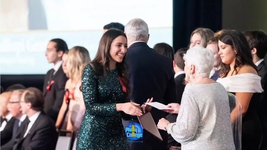 A woman in a sparkling green dress smiles as she receives a certificate from an older woman in a sparkling white dress, while a man in a suit stands between them.