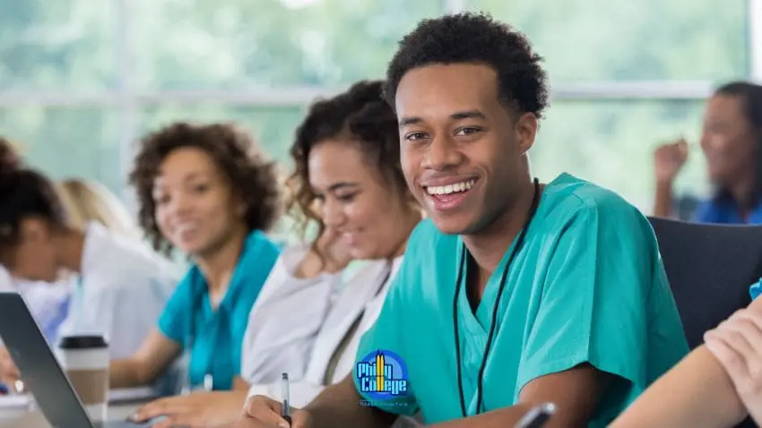 A young man in scrubs smiles at the camera while sitting at a table with other people, some wearing scrubs and others in white coats. A laptop and a coffee cup are visible on the table.