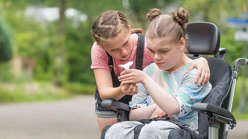 An individual in a wheelchair holds a white paper origami as another puts their arm around them.