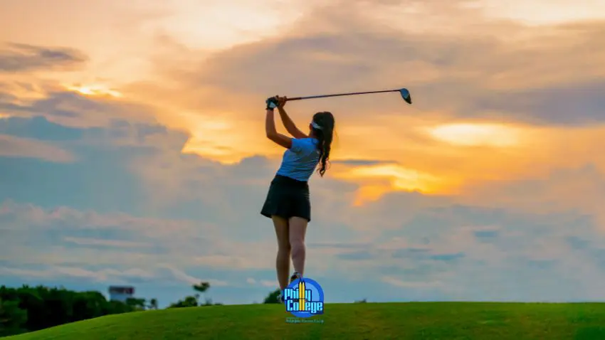 A person swings a golf club on a grassy hill against a sunset sky with orange and blue clouds.