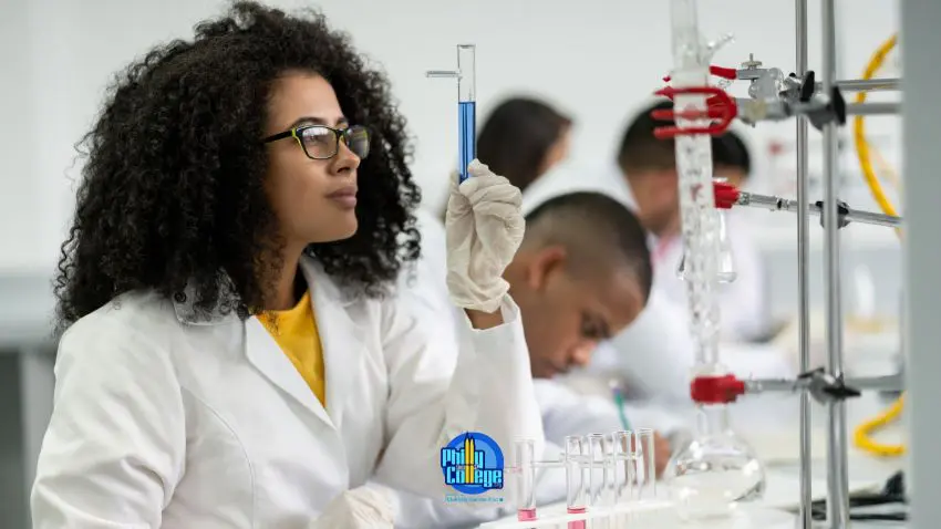 A person in a lab coat and gloves holds a test tube with a blue liquid, in a laboratory setting with other people and equipment visible in the background.